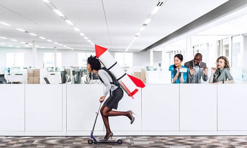 Woman speeding thru the office on the scooter with a rocket strapped to her back while her happy multiethnic  co-workers are watching her succeed in the workplace. Plenty of copyspace.